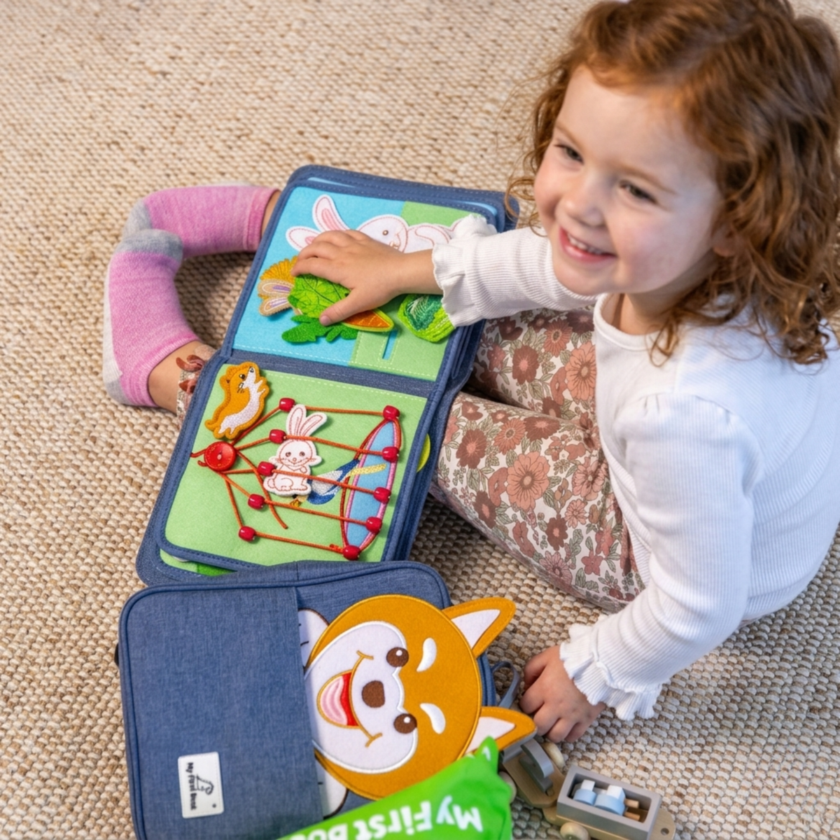 Lächelndes Mädchen spielt auf dem Teppich mit einem aufgeschlagenen Pet Friends Quiet Book – Feinmotorik mit Klett-Tieren und Schnürelementen ab 1 Jahr