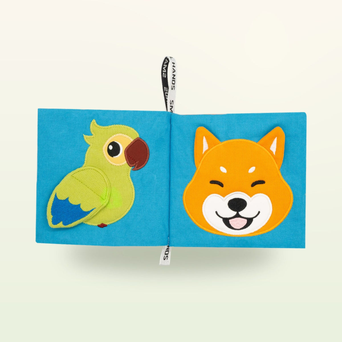 Small Hands Mascotas libro de tela Montessori abierto – loro verde y perro Shiba sonriente sobre fondo suave de color crema – a partir de 0 meses