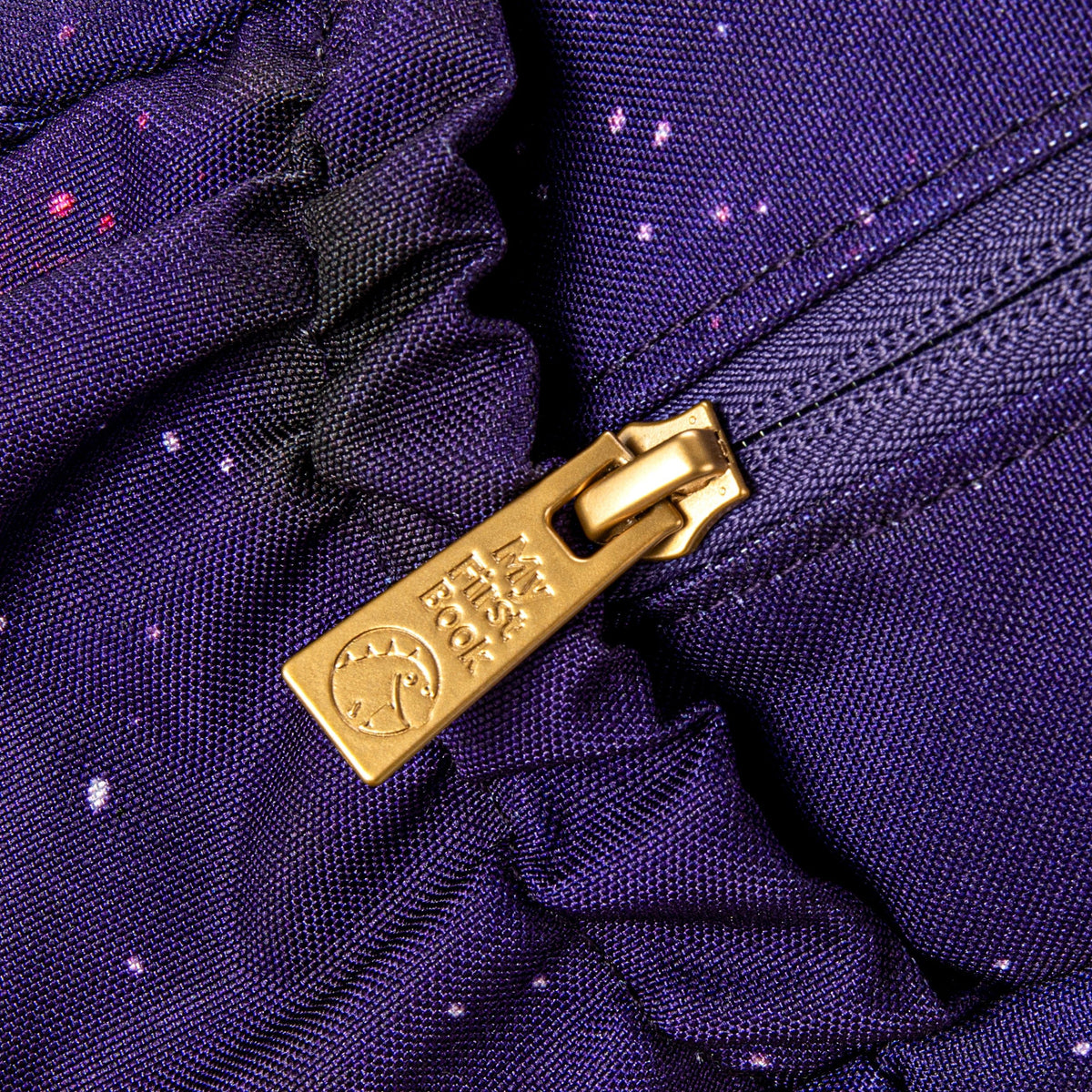 Detailaufnahme des goldenen Reißverschlusses mit -My First Book- Logo am lila Galaxie-Weltraum-Rucksack