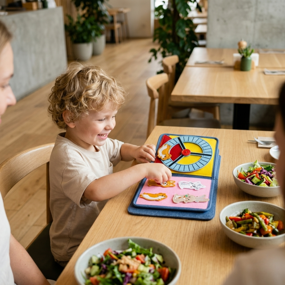 Lächelndes Kind spielt im Restaurant mit dem Pet Friends Quiet Book: zu sehen die Seite mit dem gelben Hamster-Laufrad und die Seite mit abnehmbaren Filz-Tieren wie Hund und Hase, dazu gesundes Essen in Schüsseln. Ab 1 Jahr.